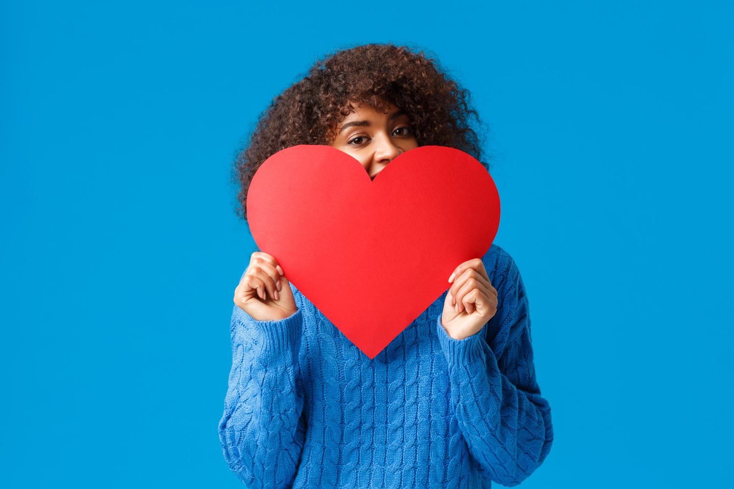 cute-and-lovely-flushed-african-american-girl-with-afro-haircut-in-sweater-hiding-face-behind-big-red-heart-and-peeking-joyfully-blue-wall