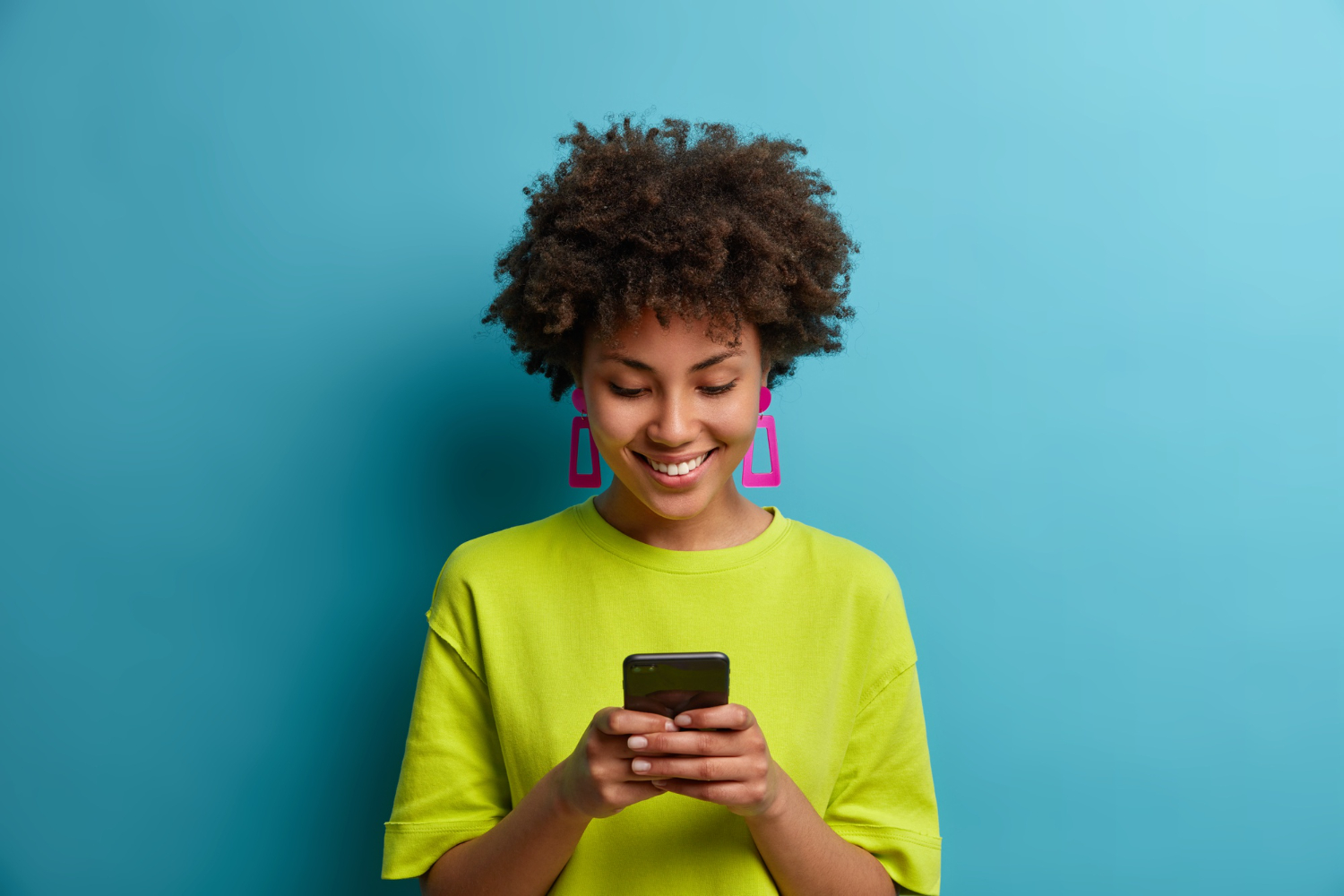 cheerful-pleased-woman-with-curly-hair-holds-mobile-phone-and-texts-with-friends-in-social-networks-uses-special-application-watches-interesting-video-isolated-on-blue-wall-people-and-technology